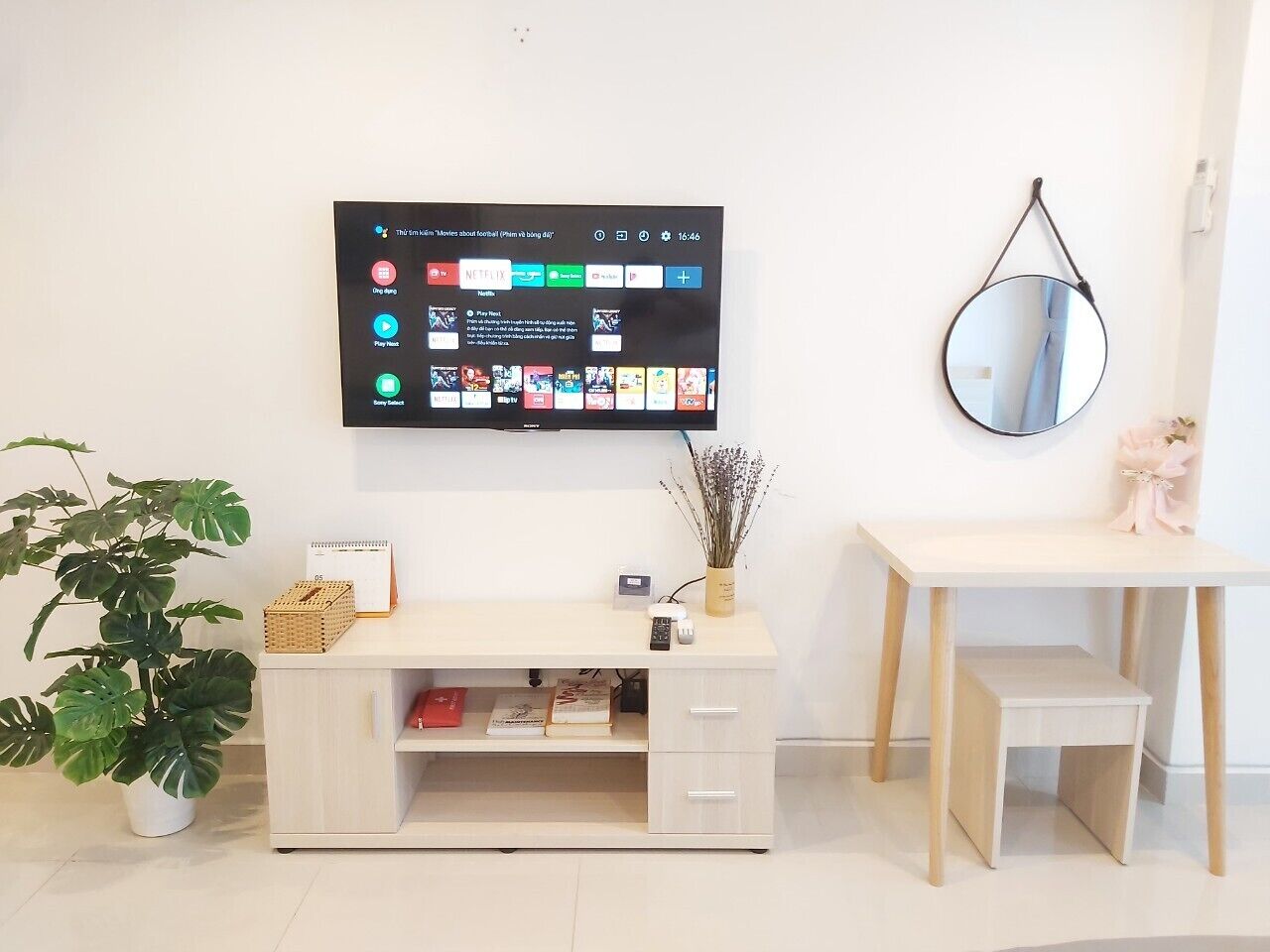 A modern living room with a flat-screen TV mounted on the wall, a wooden desk, a small table, a potted plant, and a mirror.