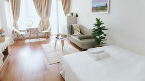 A hotel room with a bed, a sofa, a dining table, chairs, a small table with a lamp, and a large window with curtains. - Jolie Home - Sky Center - Block A (Ho Chi Minh City)
