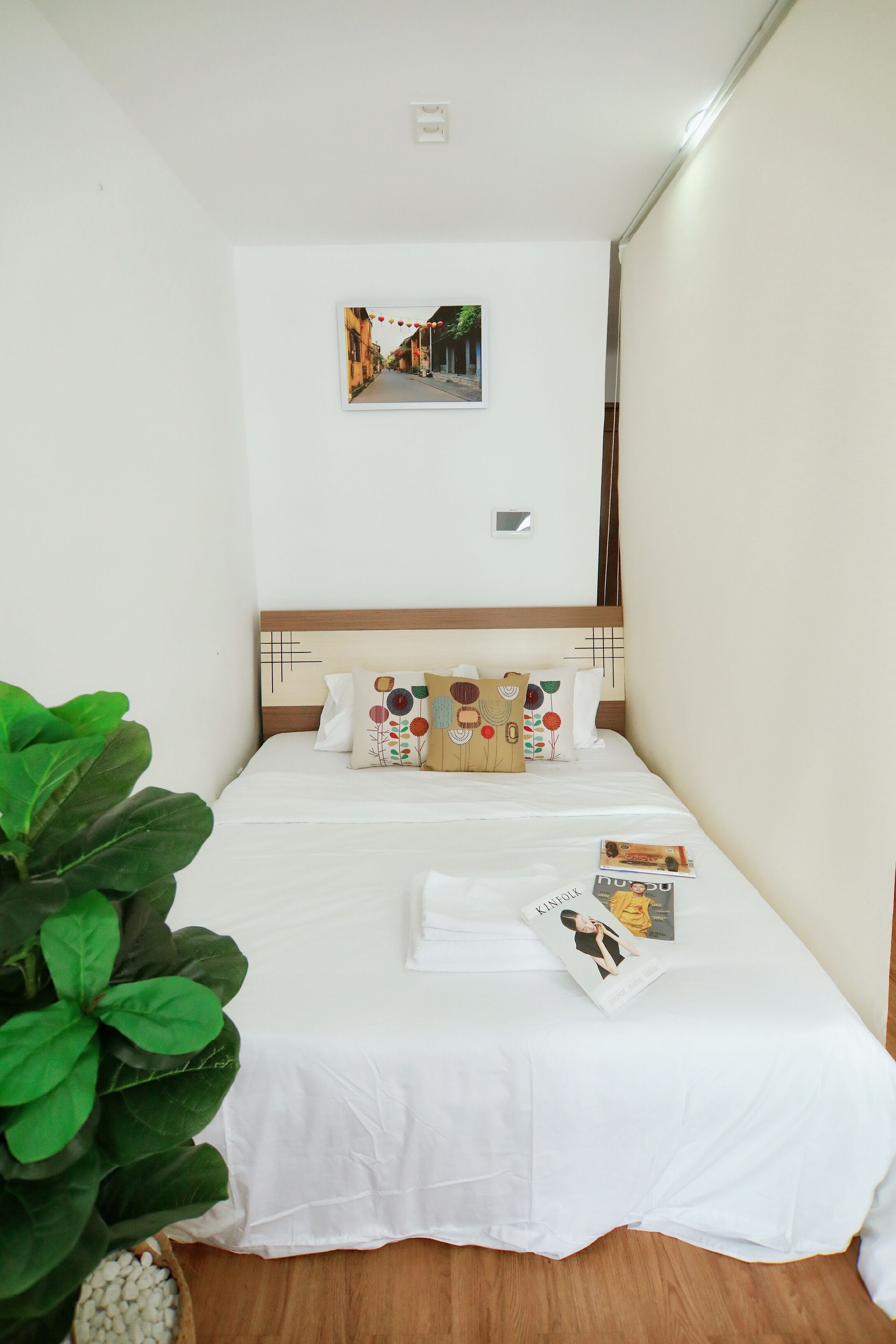 A neatly made bed with white bedding and decorative pillows, a framed picture on the wall, and a potted plant to the left.