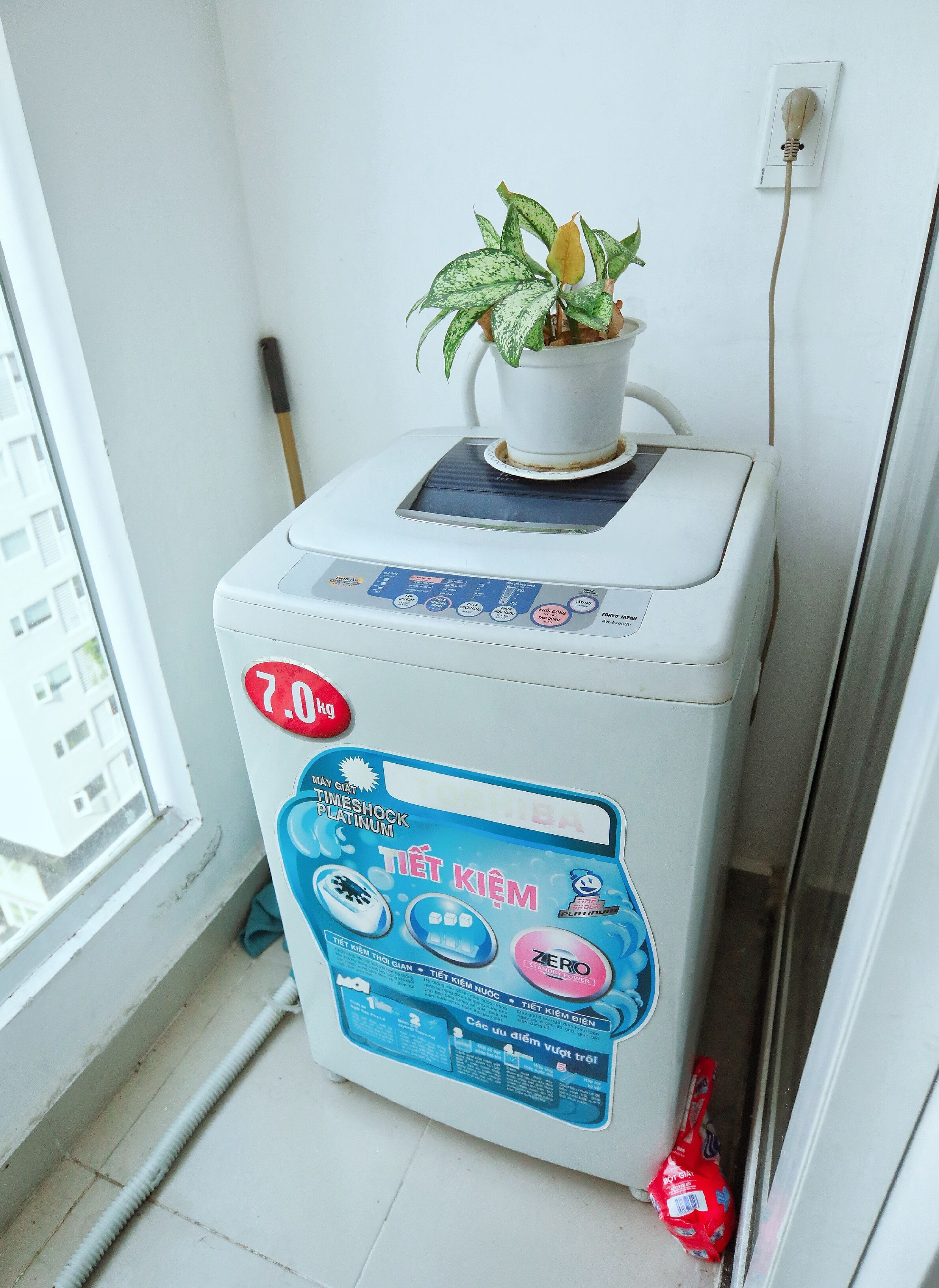 A compact, white washing machine with a control panel featuring multiple buttons and a digital display. A potted plant is placed on top of the machine, and a power cord is visible.