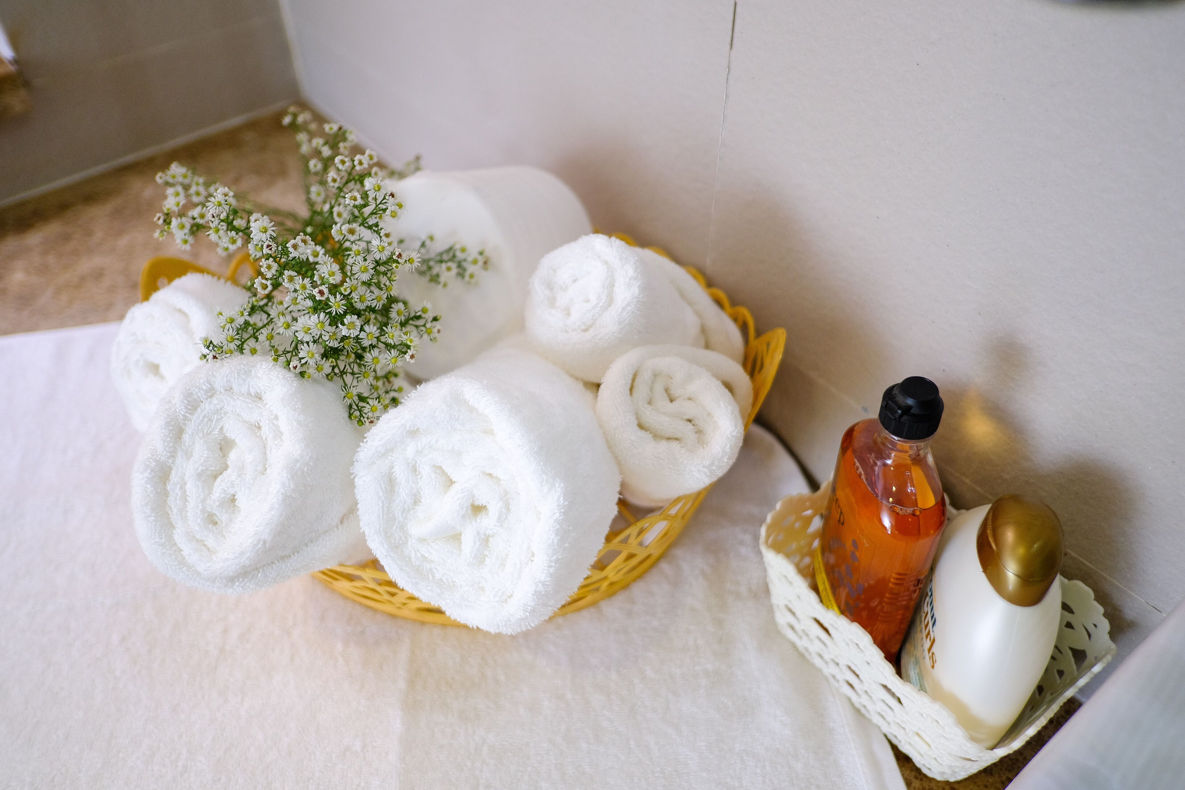 A basket of neatly rolled white towels, a bottle of orange liquid, and a white bottle with a gold cap, all placed on a white surface.