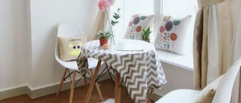 Una habitación acogedora con suelo de madera, una alfombra blanca, una mesa con mantel a cuadros, dos sillas blancas, una planta pequeña en maceta y una ventana con cortinas.