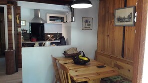 A wooden dining table with chairs, a basket of fruit, and a smartphone. A kitchen area with a microwave and sink is visible in the background.