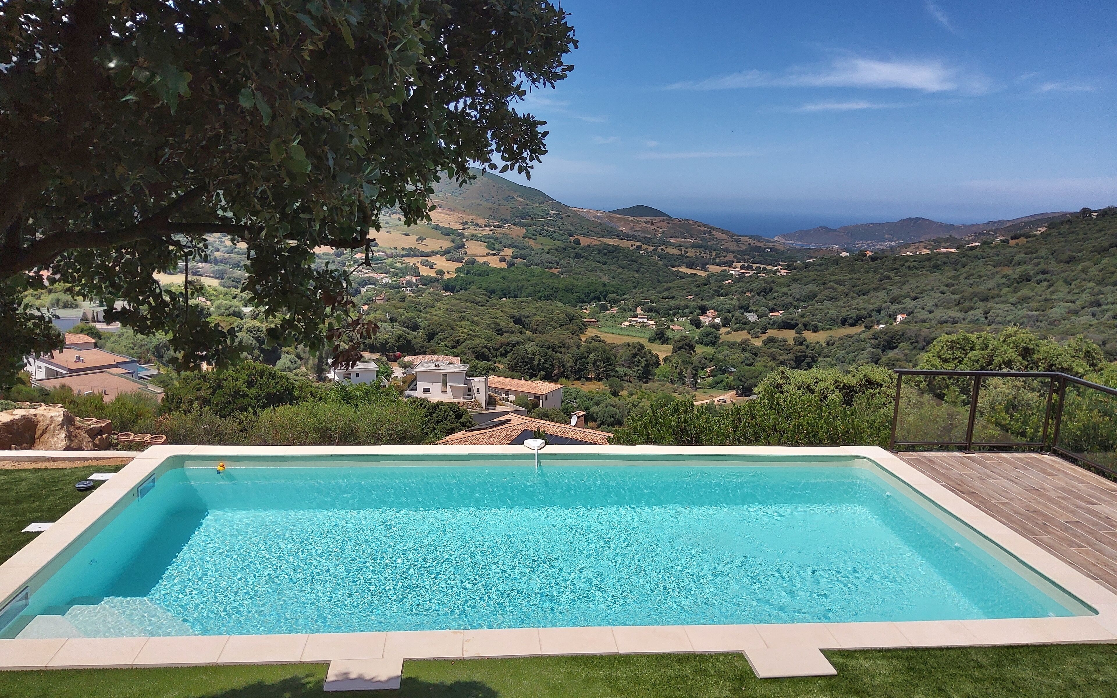 A rectangular swimming pool with clear blue water, surrounded by a wooden deck and a grassy area. In the background, there is a view of a hilly landscape with scattered houses and trees.