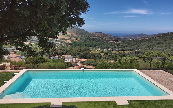 A rectangular swimming pool with clear blue water, surrounded by a wooden deck and a grassy area. In the background, there is a view of a hilly landscape with scattered houses and trees.
