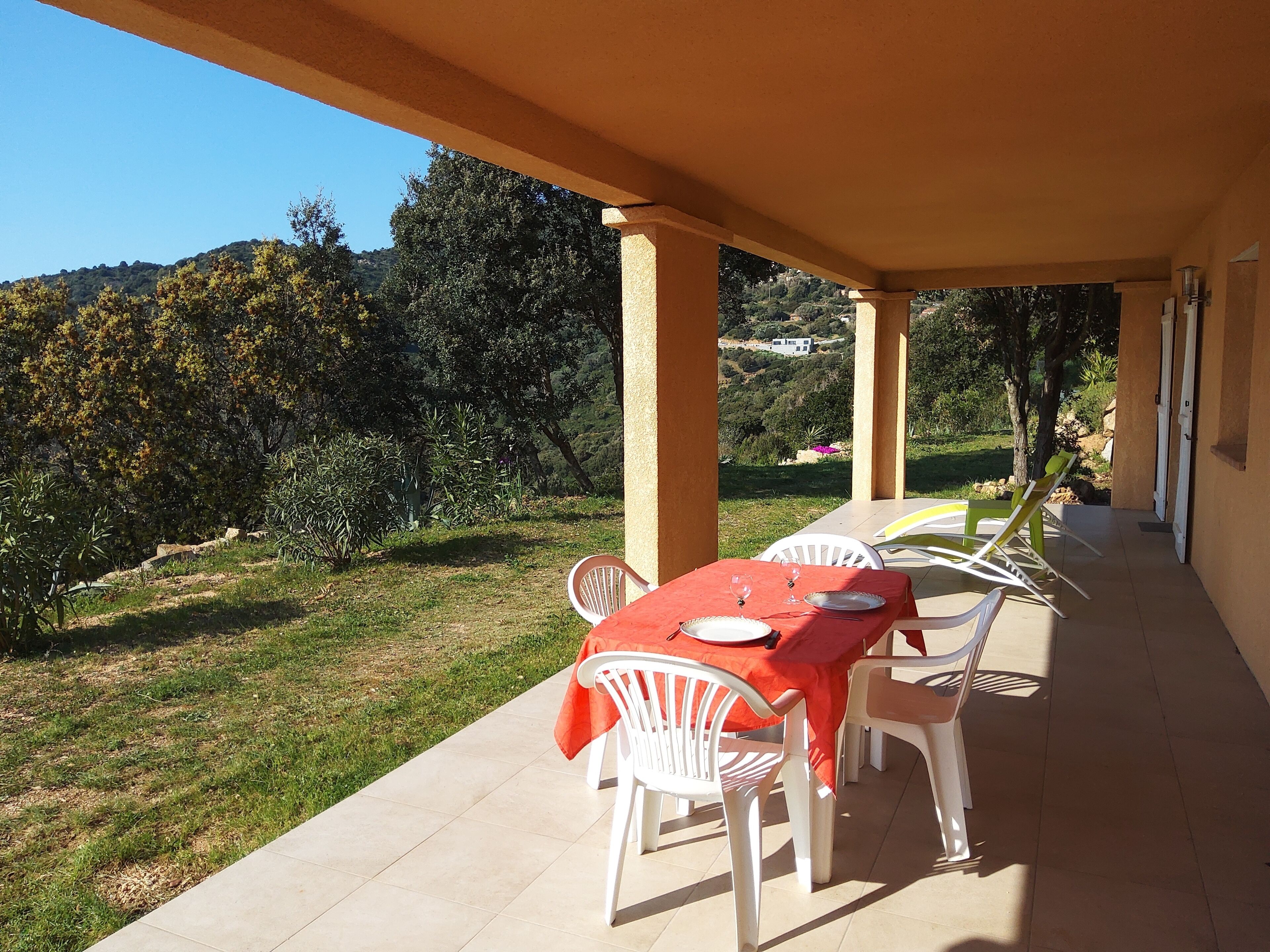 An outdoor patio with a table set for two, surrounded by white chairs, overlooking a lush garden and distant hills.