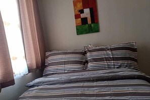 Een bed met gestreept beddengoed, een wand met een kleurrijk abstract schilderij en een raam met gordijnen.