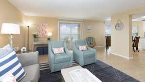 A living room with two armchairs, a sofa, a dining area, and a view through a large window.