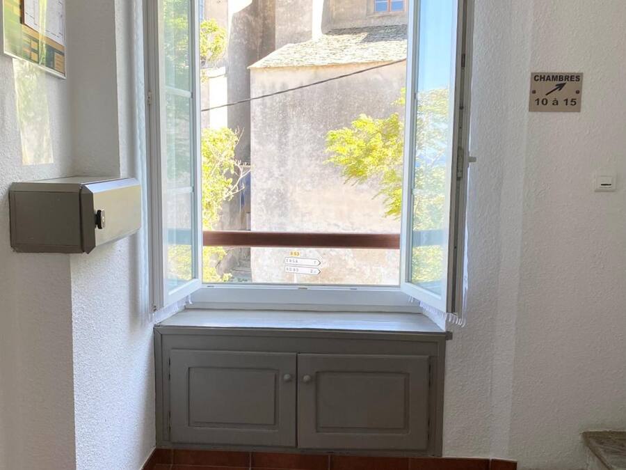 Una habitación con ventana a un patio interior, un banco integrado y una puerta con un cartel que indica el número de habitación 10 al 15.