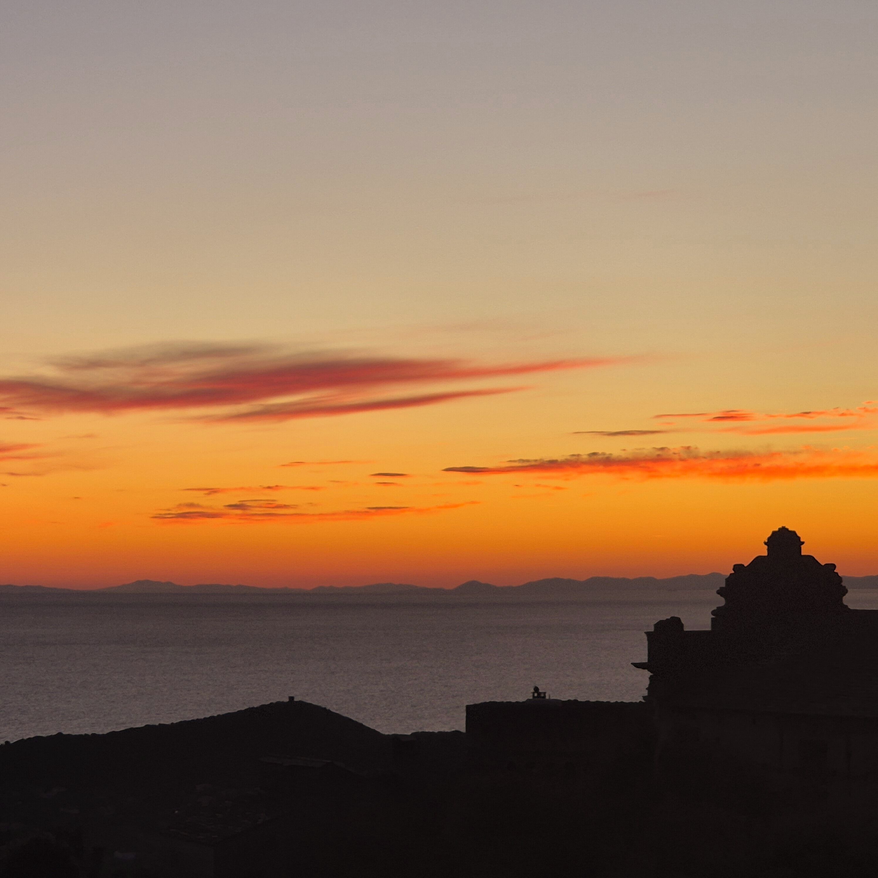 A coastal sunset with silhouettes of rock formations and distant mountains.