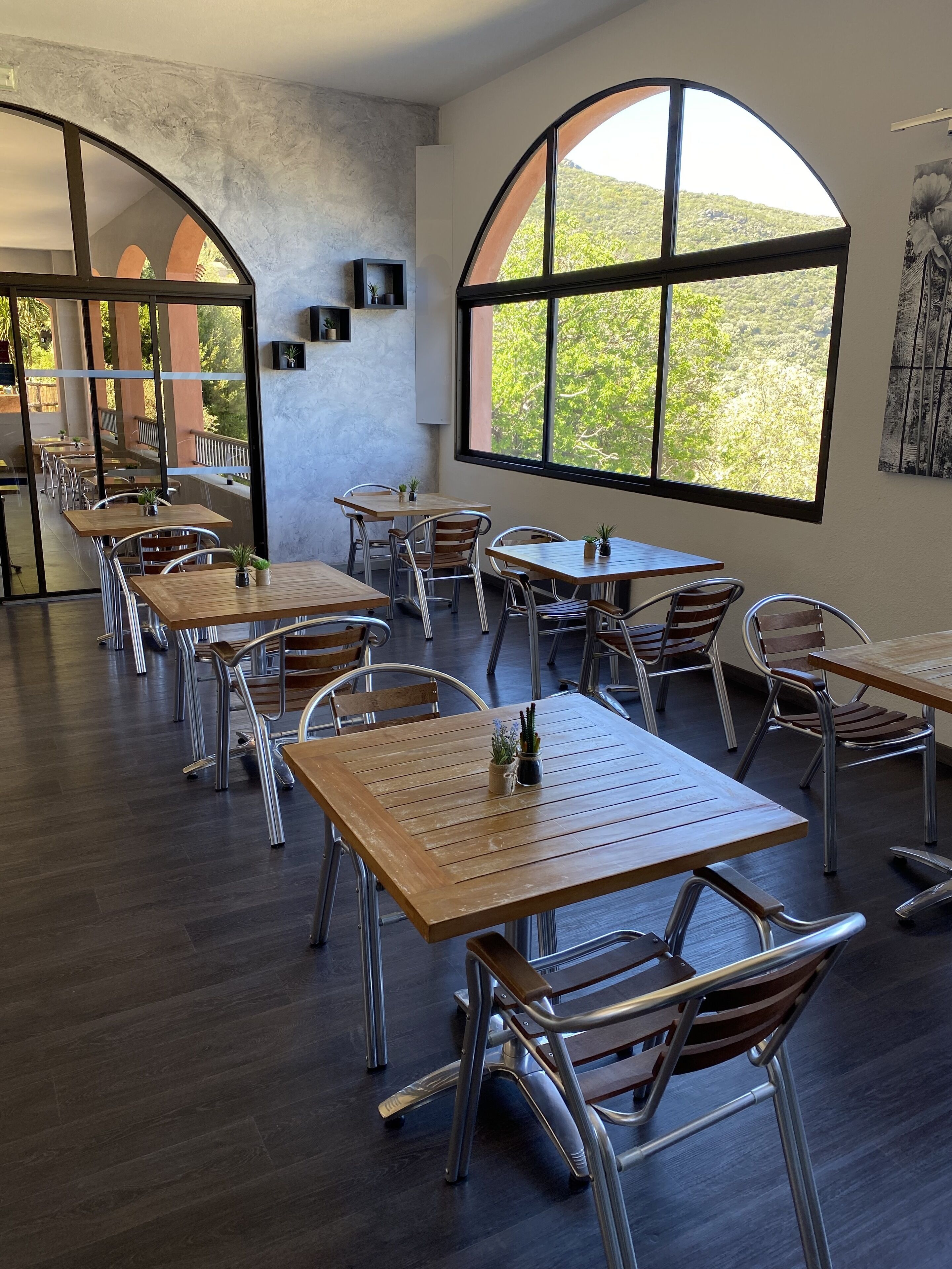 A modern dining area with wooden tables and metal chairs, large arched windows offering a view of greenery, and a concrete wall with a shelf.
