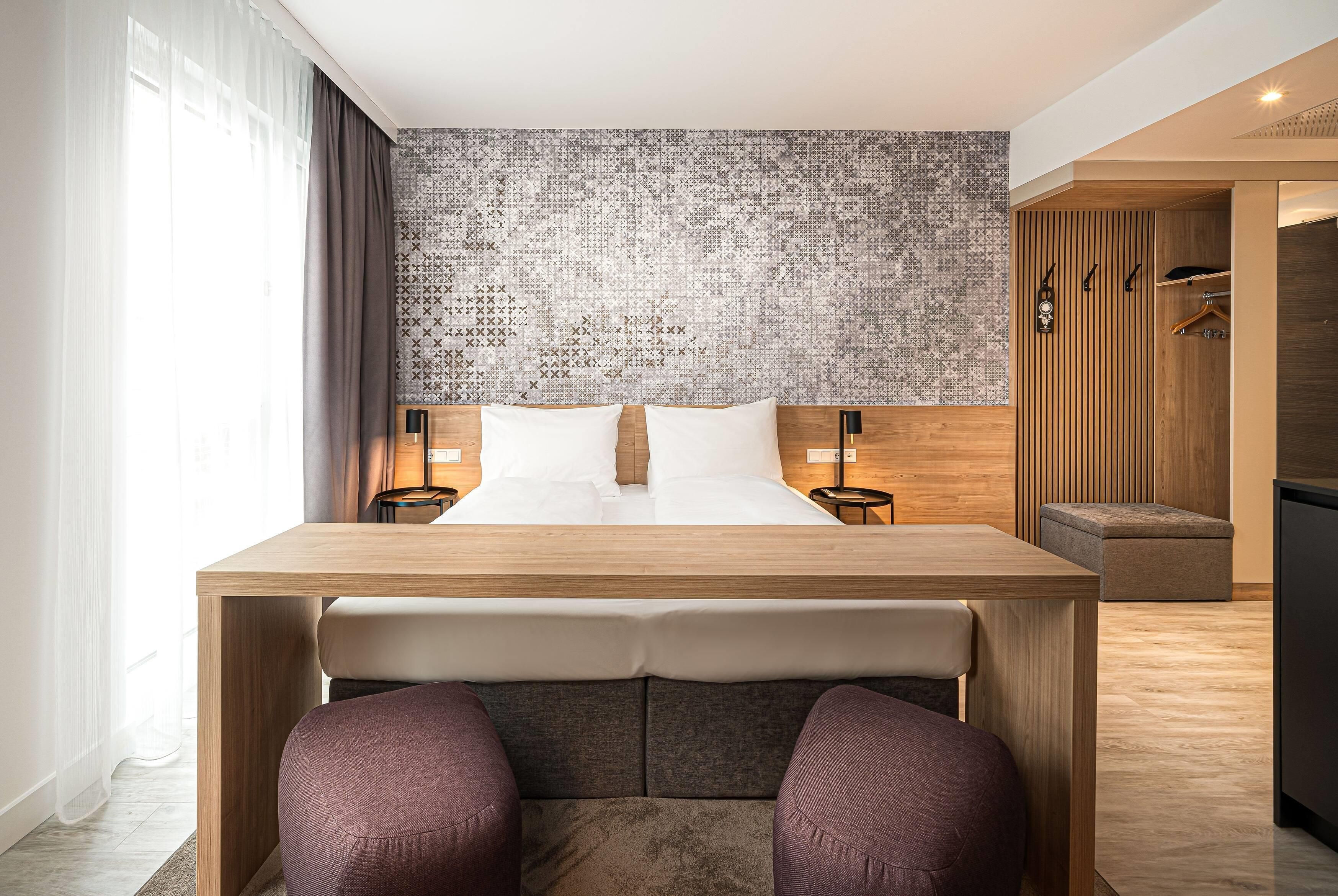 Una habitación de hotel moderna con una cama de madera, una mesita, dos sillas, un papel tapiz decorativo y un armario con espejo.