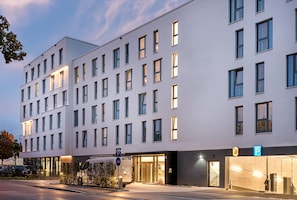 Un hotel de varios pisos con fachada moderna, numerosas ventanas y un estacionamiento con un cartel que indica un lugar disponible.