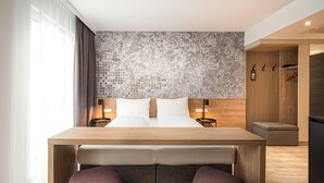 Une chambre d’hôtel moderne avec un lit en bois, une petite table, deux chaises, un mur à motifs et une armoire avec un miroir.