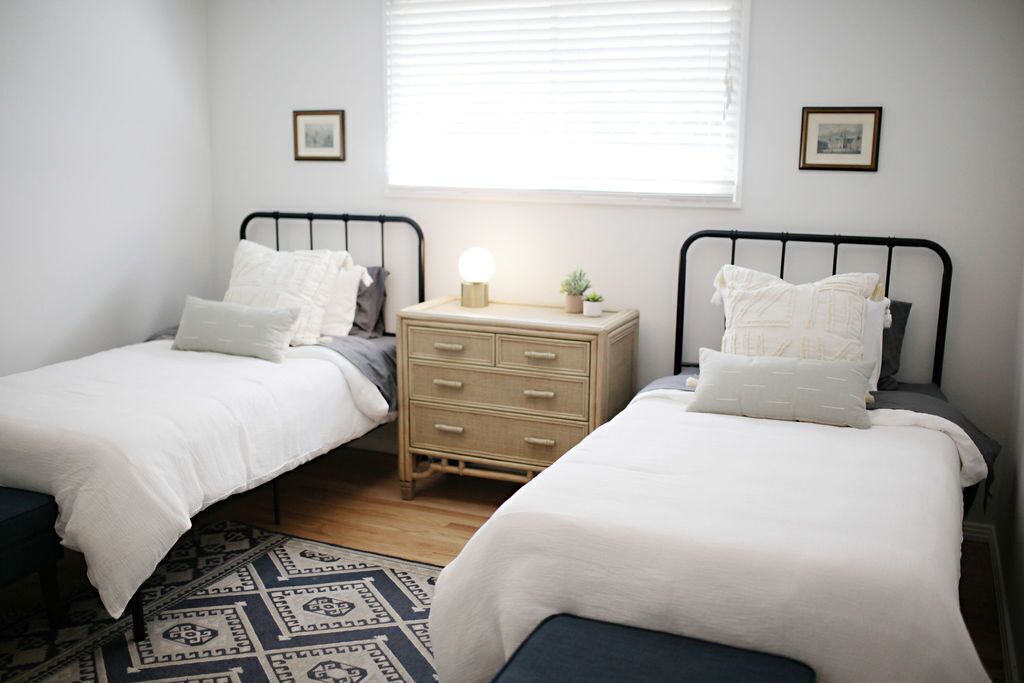 A bedroom with two single beds, a bedside table with a lamp, and a window with blinds.