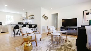 Una sala de estar moderna con una cocina al fondo, que incluye un televisor de pantalla plana, zona de estar con sillas y una mesa de centro, y una alfombra con estampado de cebra.