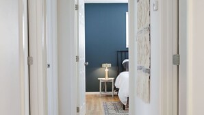 Un pasillo con suelo de madera, paredes blancas y una puerta que da a una habitación con cama y una mesita de noche con lámpara.