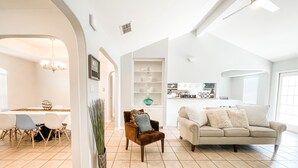 A spacious living room with a high ceiling, a dining area with a white table and chairs, and a kitchen in the background.