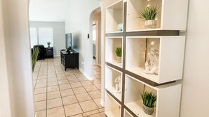 A hallway with a white tiled floor, a white wall with a built-in shelf unit, and a doorway leading to another room.