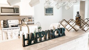 A kitchen with a white brick backsplash, stainless steel appliances, hanging mugs, and a 'HAPPY HOUR' sign.