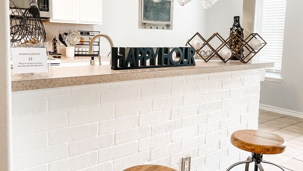 A kitchen with a white brick backsplash, a bar area with stools, hanging mugs, and a 'HAPPY HOUR' sign.