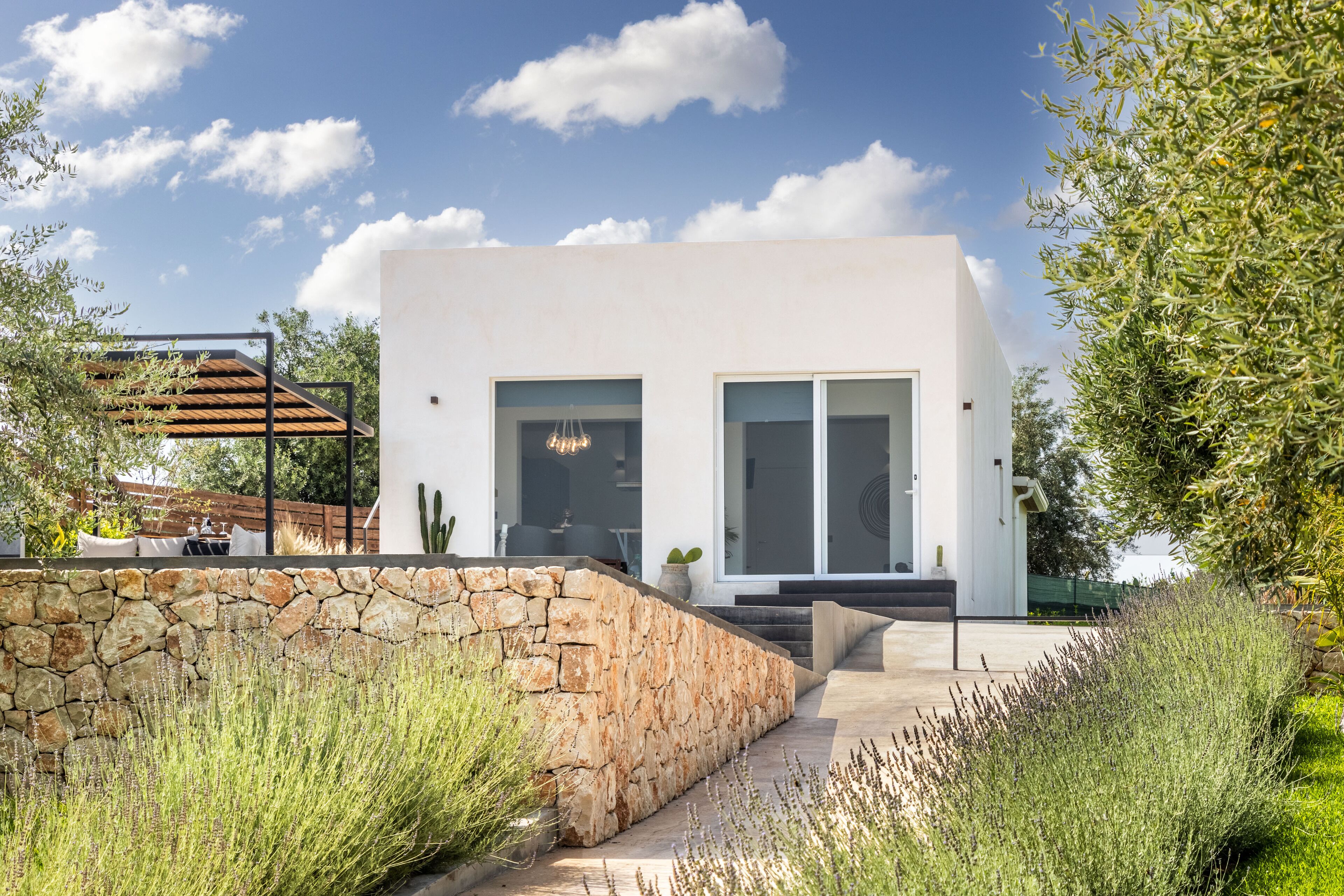 Una casa moderna con pared de piedra, pérgola de madera y jardín con diversas plantas.