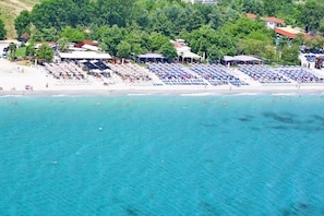 An aerial view of a beach with rows of sun loungers and umbrellas, a sandy shore, and a backdrop of trees and buildings.