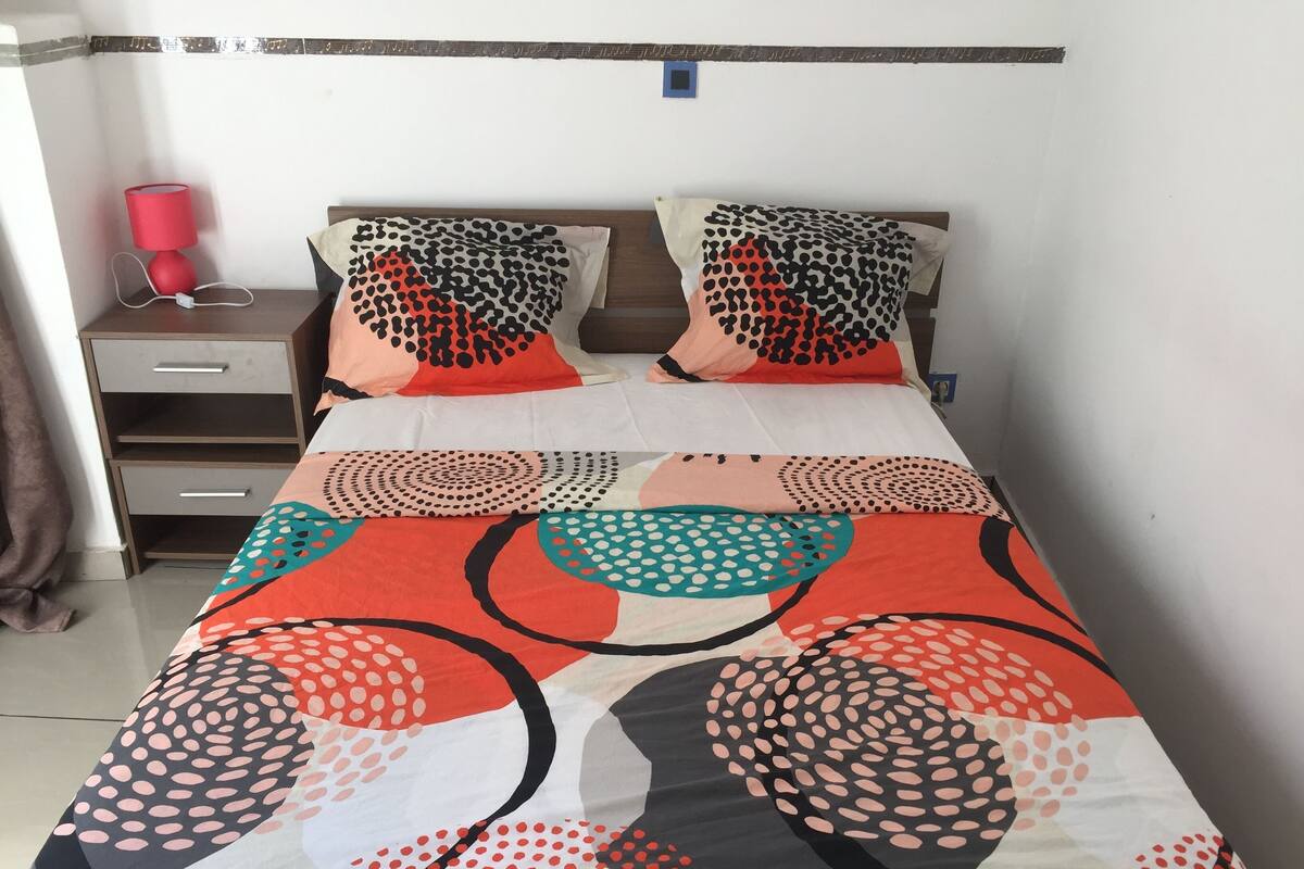 Una cama con una colcha estampada, dos almohadas con diseño de lunares, una mesita de noche con una lámpara roja y un mueble con cajones.