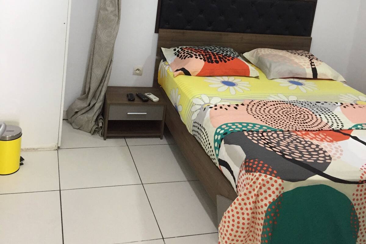 Un dormitorio con una cama, una mesita de noche, un ventanal con cortinas, un aire acondicionado instalado en la pared y un cubo de basura amarillo.