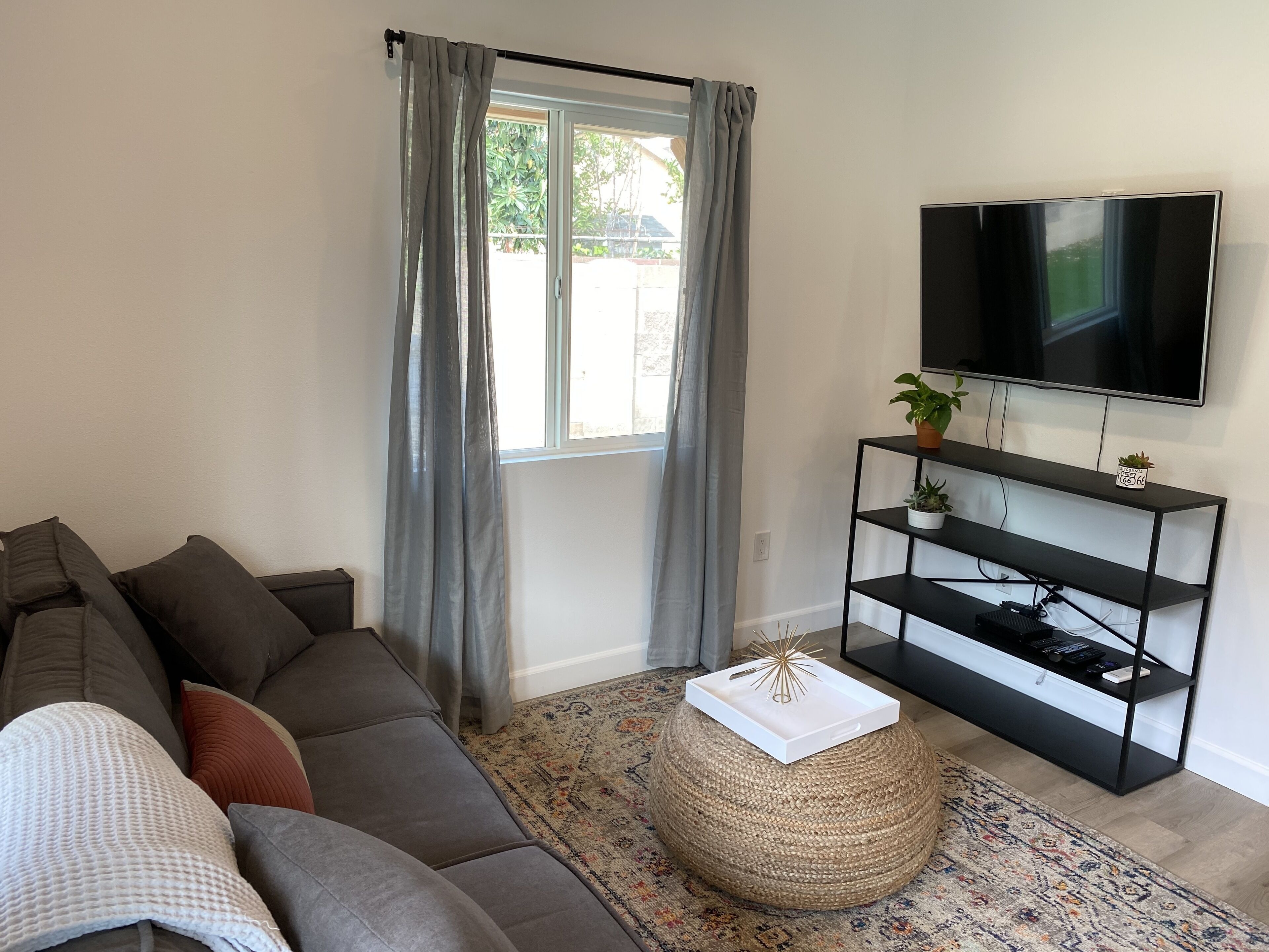 A living room with a grey sofa, a TV on a black stand, a woven ottoman, and a patterned rug.
