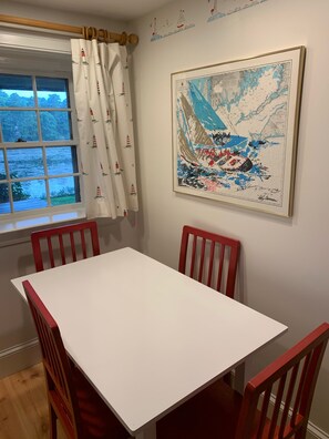Un coin repas avec une table blanche et des chaises rouges, une fenêtre avec des rideaux et une carte encadrée accrochée au mur.