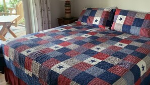 Une chambre avec un grand lit, une table de chevet avec une lampe, un tableau encadré au mur et une vue sur l’extérieur par une fenêtre.