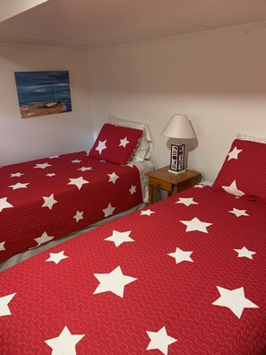 Une chambre avec deux lits, chacun recouvert d’une literie rouge ornée d’étoiles blanches, une petite table de chevet en bois avec une lampe, et un tableau représentant un bateau accroché au mur.