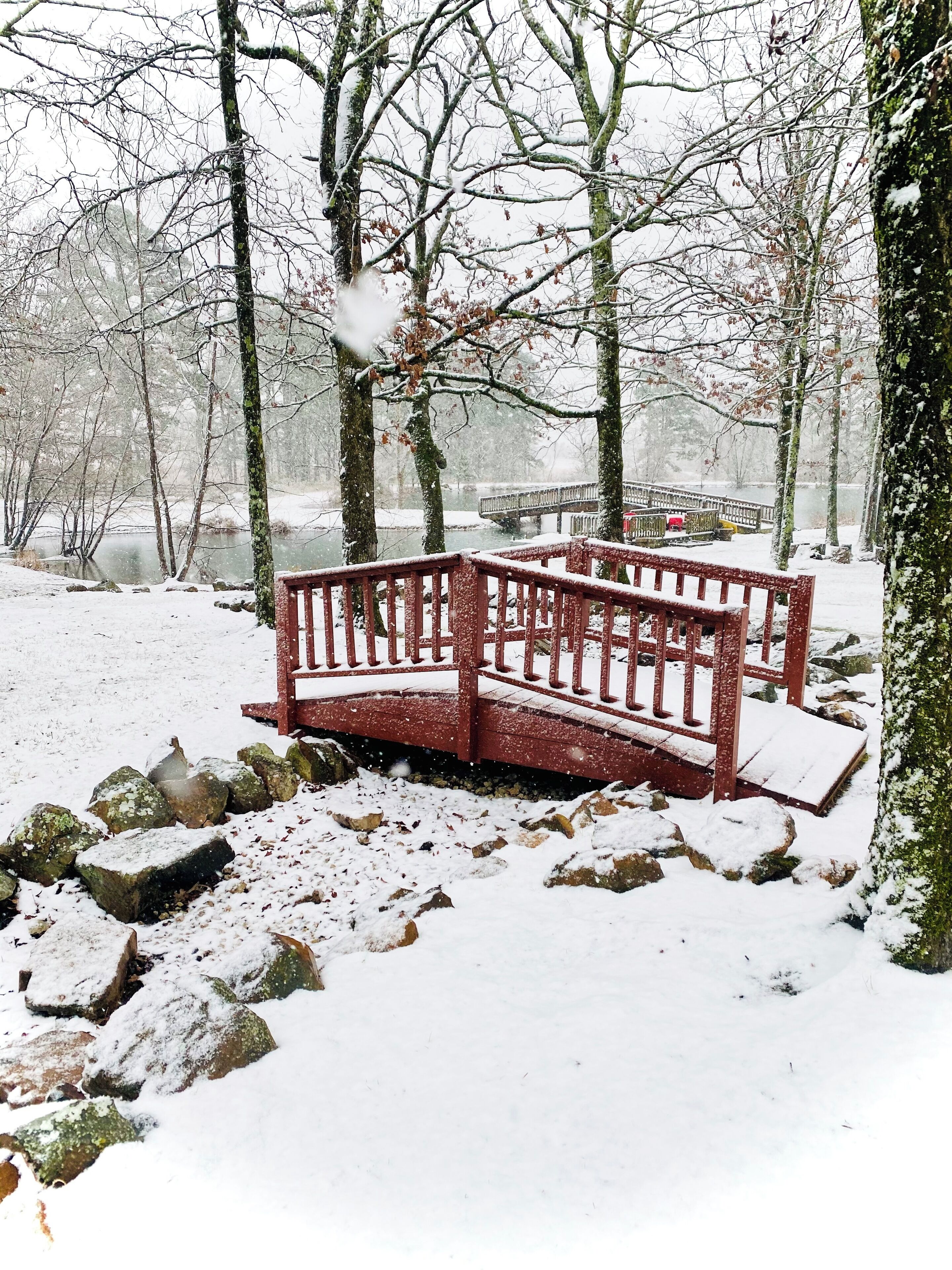 A snow-covered bench in a park with a bridge in the background.