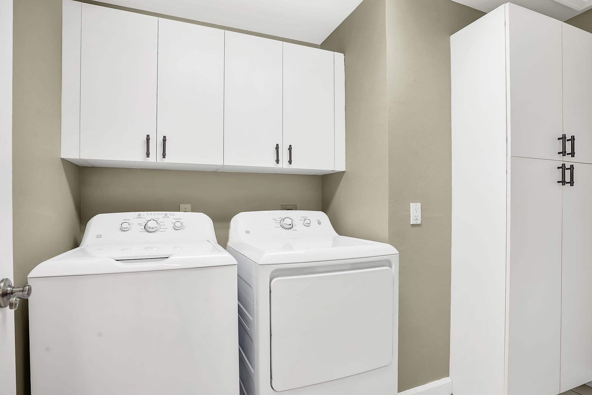 A laundry room with two white washing machines, a cabinet above, and a tall white wardrobe.