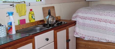 A small kitchen with a sink, stove, and wooden cabinets. There are cleaning supplies on the counter and a window with a view of greenery outside.