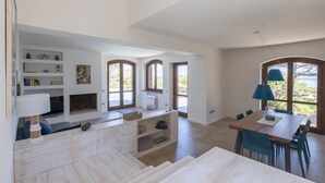 Uma sala de estar ampla com escada de madeira, mesa de jantar com cadeiras e área de cozinha com prateleiras embutidas e lareira.