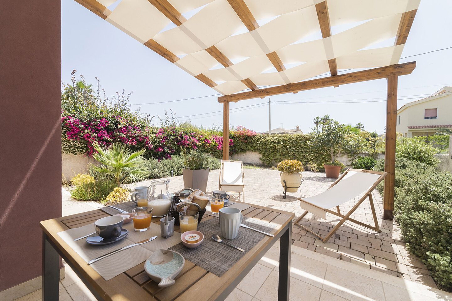 Une terrasse extérieure avec une table dressée pour le petit-déjeuner, entourée de chaises et d’une pergola.