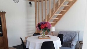 A dining area with a round table, chairs, a vase with flowers, a wooden staircase, and a blue sofa. - 2-person gîte 7 minutes from surf spots, beaches and hiking trails. (PLONEOUR LANVERN)