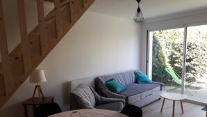 A modern living room with a grey sofa, teal cushions, a small round table with a white tablecloth, and a wicker basket. - 2-person gîte 7 minutes from surf spots, beaches and hiking trails. (PLONEOUR LANVERN)