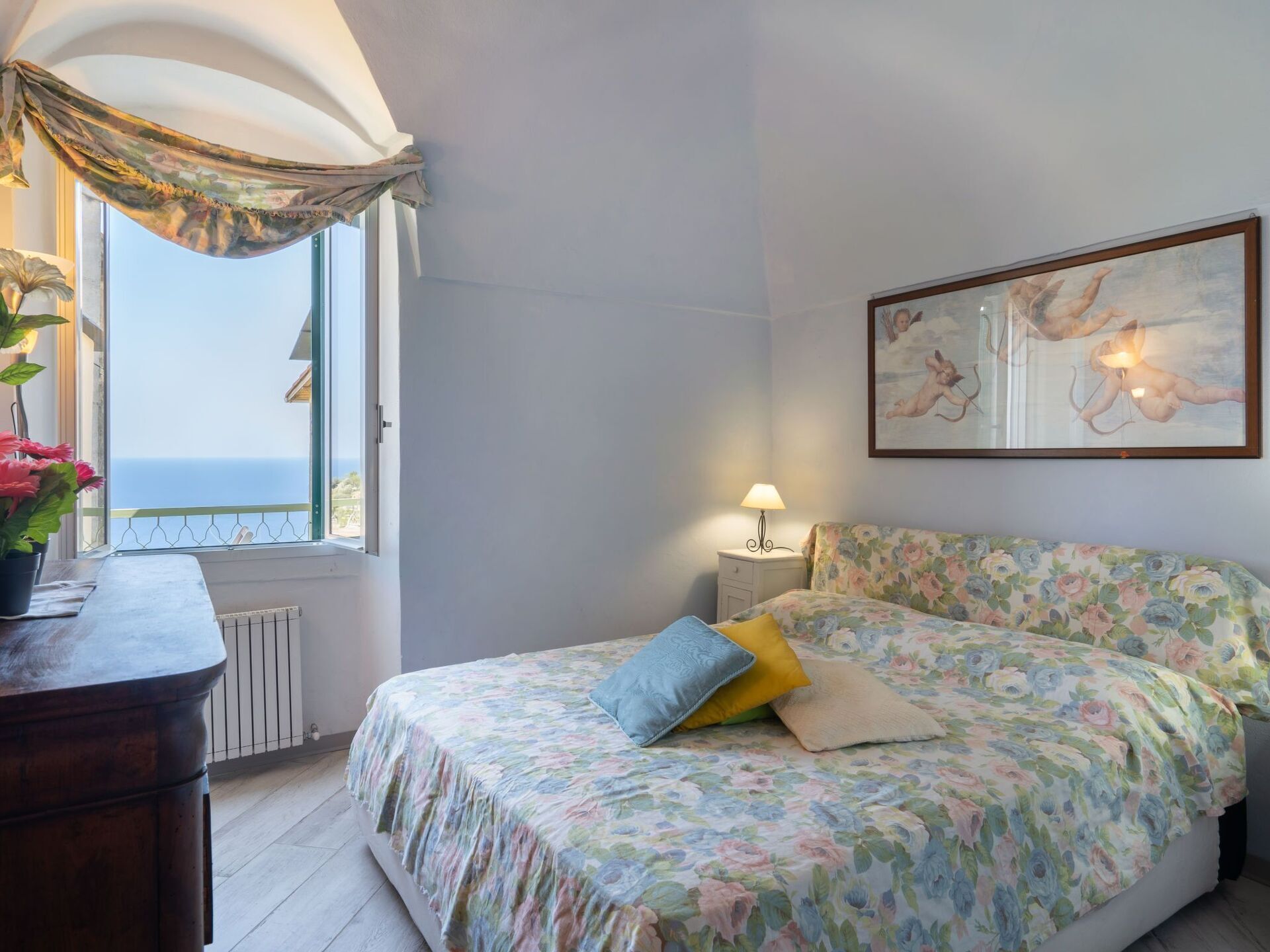 A bedroom with a floral bedspread, a nightstand with a lamp, a painting on the wall, and a view of the sea through a window.