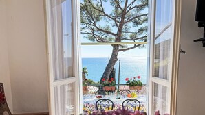 Une fenêtre ouverte donnant sur une table dressée pour le repas, une chaise et un pot de fleurs.