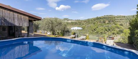 A swimming pool with a view of a hillside, a building with a stone facade, and a white umbrella.