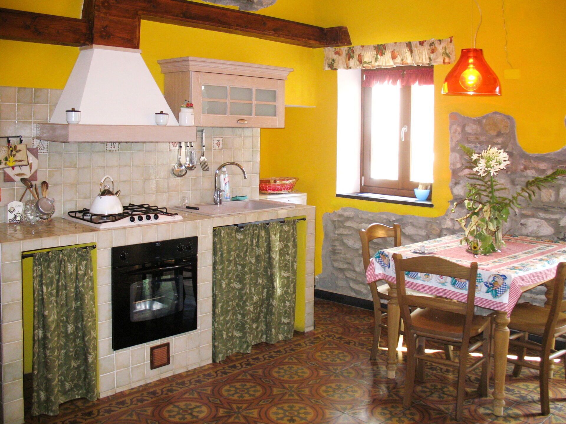 Eine Küche mit Gasherd, Backofen und Spüle, ein Esstisch mit Stühlen und ein Fenster mit Vorhang.