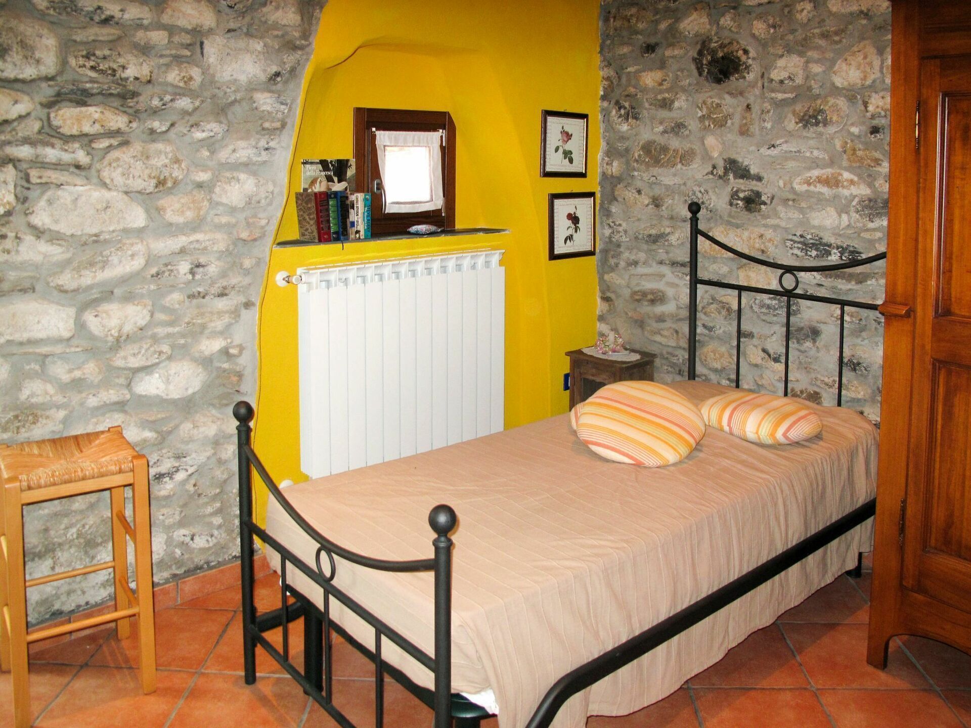Un dormitorio con pared de piedra, pared amarilla, cama con almohadas a rayas, una silla de madera, un radiador y una mesita con un jarrón de flores.