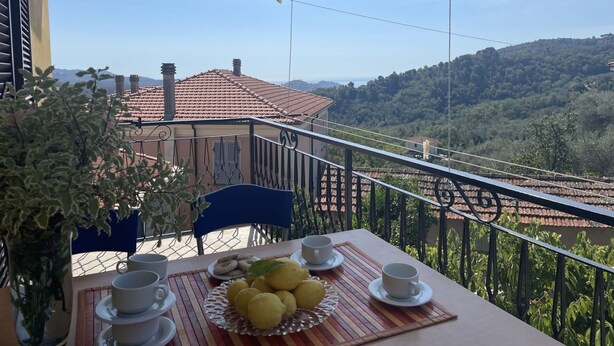 Varanda com mesa posta para dois, com vista para uma paisagem cênica com um edifício de telhado vermelho e vegetação exuberante.