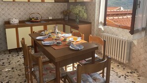 Uma área de refeições com uma mesa de madeira posta para o café da manhã, rodeada por cadeiras de madeira. Uma cozinha com armários amarelos e uma janela com vista para o campo é visível.