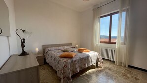 Quarto com cama, mesa de cabeceira, escrivaninha, luminária e janela com vista.