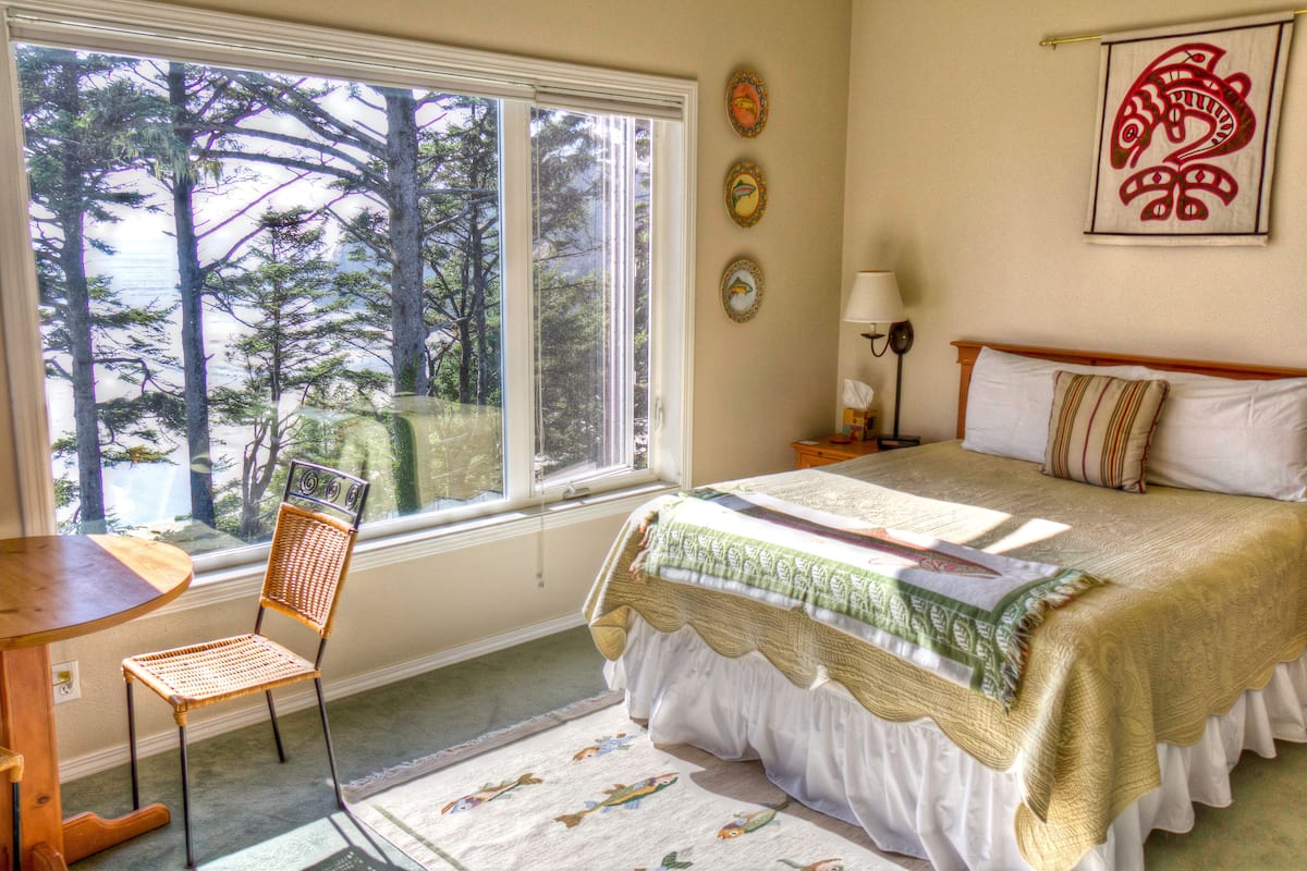 A bedroom with a large window, a bed with a green and white bedspread, a chair, and a nightstand with a lamp.