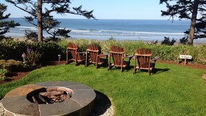 A circular fire pit surrounded by six wooden Adirondack chairs facing a beach. - Tyee Lodge Oceanfront B&B (Newport)